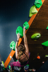 A young, slender, athletic climber in a sports field climbs a boulder at a festival in a climbing center. A vertical frame.