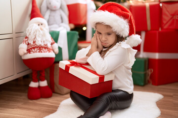 Adorable hispanic girl holding gift sitting on floor by christmas tree with sad expression at home