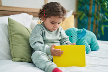 Adorable hispanic girl reading book sitting on bed at bedroom