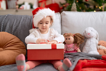 Fototapeta na wymiar Adorable caucasian girl holding gift sitting on sofa by christmas tree at home