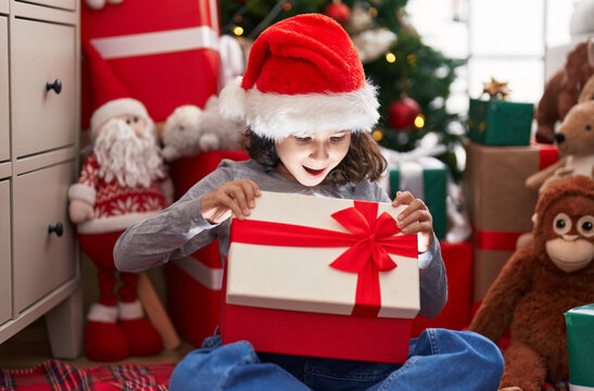 Adorable chinese girl unpacking gift with surprise expression sitting by christmas tree at home