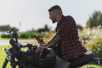 Tattooed Caucasian man wearing a red plaid shirt looking at his phone while on a motorcycle