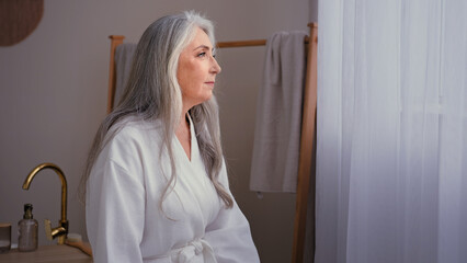 Pensive aged elderly lady 60s old Caucasian 50s senior mature gorgeous woman in white bathrobe standing in bathroom at home. Calm grandmother female with gray hair moisturizing rejuvenating skin care