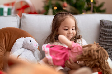 Obraz na płótnie Canvas Adorable hispanic girl holding doll sitting on sofa by christmas tree at home