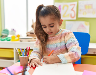 Adorable hispanic girl student sitting on table drawing on paper at kindergarten