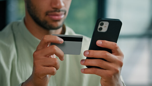 African American bearded man indoors unrecognizable male customer holding credit card and smartphone sitting at home office using easy mobile payment app service making purchase buying in online store