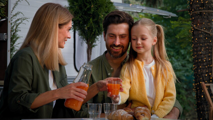 Caucasian family on camping vacation outdoors sitting near van have lunch picnic eat good with croissants mother pouring fresh orange juice into glasses for father husband and child little daughter
