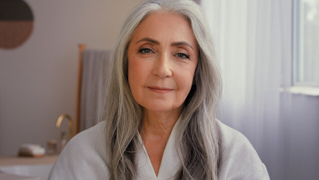 Portrait indoors old Caucasian 50s senior model gray-haired mature woman 60s lady in bathroom showing jar of anti-aging cosmetic natural organic sunscreen mineral cream moisturizing face skin care