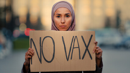 Portrait female muslim islamic girl woman in hijab standing in city outdoors showing cardboard banner sign text no vax protest against immunization covid19 coronavirus vaccination vaccine denial