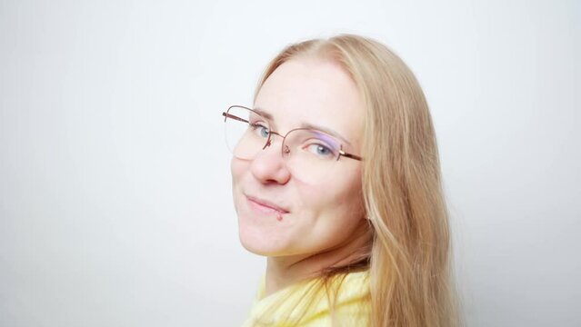 Close-up portrait young beautiful blonde woman in glasses and casual yellow sweater turns around, wave her hair, and looks at the camera with a
cute charming look. White studio background stock video
