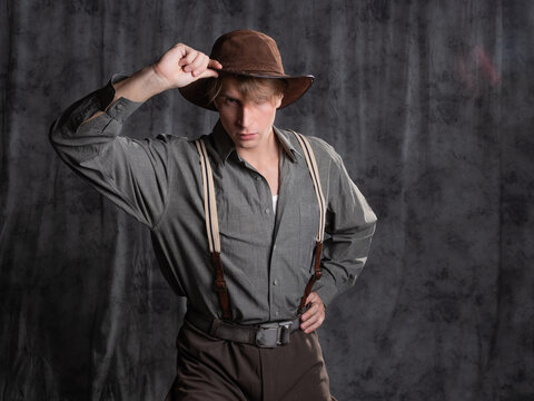 A young man in retro style, an adventure character. A guy in a hat and a gray shirt, breeches with suspenders.