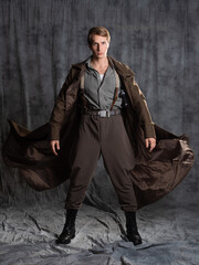 hero in a military style with a gun in his hands, a young man in a long brown coat and breeches with suspenders. Photo in the studio on a gray background - 551118175