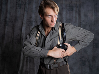 Fototapeta premium A lawyer with a pistol in a holster, gray shirt and trousers with suspenders, retro style. A young man poses in the studio on a gray background