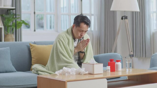 Sick Asian Man With Blanket Trying To Get Warm On Sofa In The Living Room At Home
