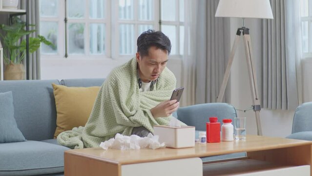 Sick Asian Man With Blanket Using Smartphone On Sofa In The Living Room At Home
