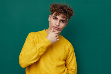 Obraz na płótnie Canvas a cute, thoughtful man stands on a green background in a yellow sweater holding his hand to the will of the face. Horizontal photo with empty space