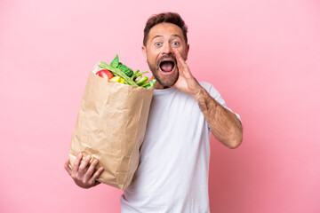 Middle age man holding a grocery shopping bag isolated on pink background with surprise and shocked...