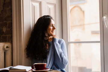 Attractive girl warms herself with hot coffee in a cafe in cold winter. Young woman with cup of coffee relaxing in restaurant and looking out the window.