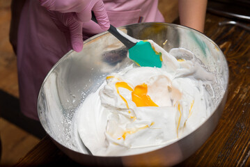 Stirring the egg yolk into the whipped whites with an acrylic spatula in a metal bowl - obrazy, fototapety, plakaty