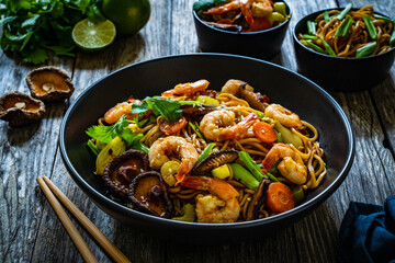 Mie noodles with fried prawns, shiitake mushrooms, coriander, carrot and celery on wooden table
