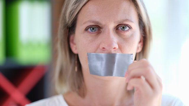 Female with serious face removes adhesive tape from her mouth close up.