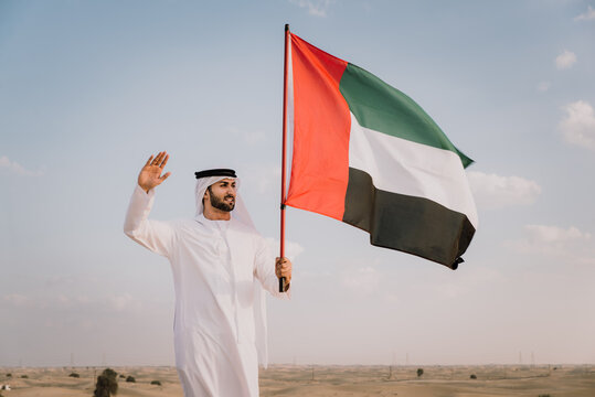 Arab man wearing typical middle eastern clothing in the desert and holding the emirate flag