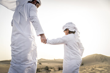 Playful arab man and his son wering traditional middle eastern emirate clothing playing and having fun in the desert of Dubai - obrazy, fototapety, plakaty