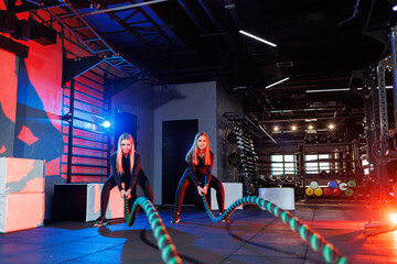 Fototapeta premium Two young women exercising with ropes ropes in the gym. Woman doing rope workout together with trainer