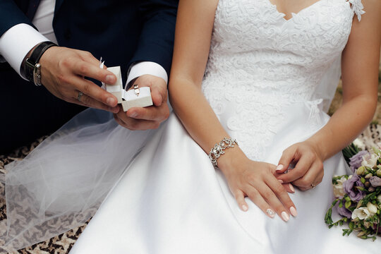 A groom in a dark blue suit offers to take a gold wedding ring from a box to his bride in a white dress. Image for your creativity, design or illustrations.