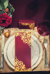 Composition on the table from table setting, fork, knife, glass, white plate with a holiday card in red and gold colors. The table is decorated with flowers and golden walnuts.