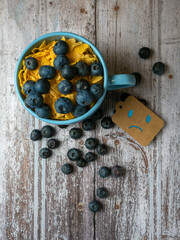 Close-up of a blue mug with a sad face drawn on it filled with blueberries and cereal, blue monday...