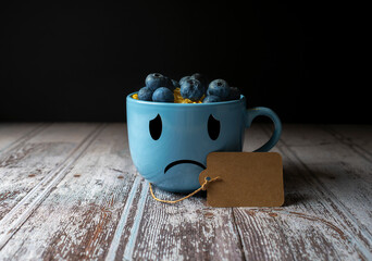 Close-up of a blue mug with a sad face drawn on it filled with blueberries and cereal, blue monday...