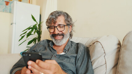 Middle-aged man relaxing on his couch while in home and using his smartphone to text