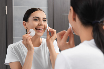 Young woman using cotton pads with micellar water near mirror in bathroom