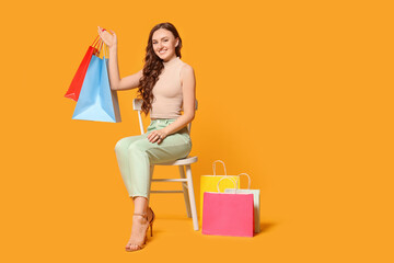 Happy woman holding colorful shopping bags on chair against orange background