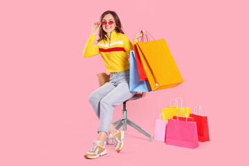 Happy woman in stylish sunglasses holding many colorful shopping bags on armchair against pink background