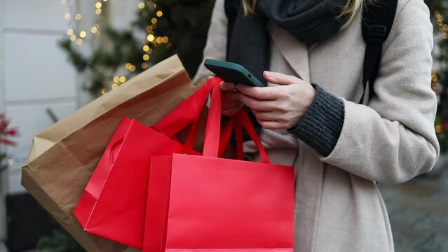Cropped image of female hands holding red holiday gifts packages used phone on the city street near the shopping center. Retail sale concept. Shopper. Sales. Shopping Center.Cyber Monday.Black Friday