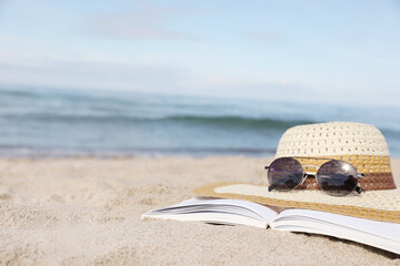 Open book, sunglasses and hat on sandy beach near sea, closeup. Space for text