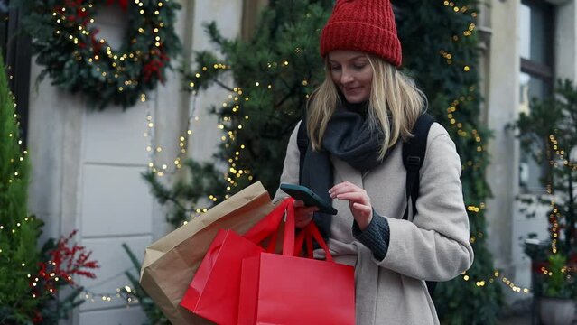Cropped image of female hands holding red holiday gifts packages used phone on the city street near the shopping center. Retail sale concept. Shopper. Sales. Shopping Center.Cyber Monday.Black Friday