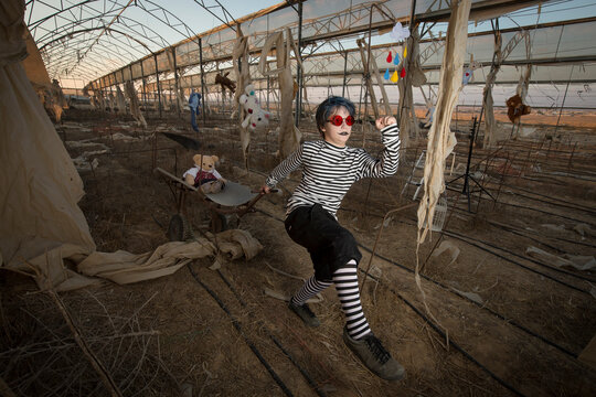 a boy in red sunglasses, a scary smile in a clown costume and make-up plays with an old cart, in an abandoned greenhouse among hanging toys