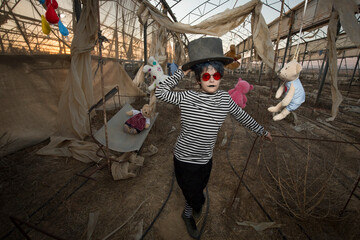 a boy in red sunglasses, a scary smile in a clown costume and make-up plays with an old cart, in an abandoned greenhouse among hanging toys