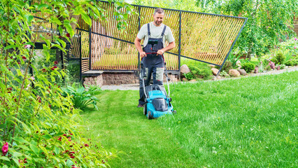 A man mows the grass with an electric lawn mower on a hot summer day. Gardener with a lawn mower in...