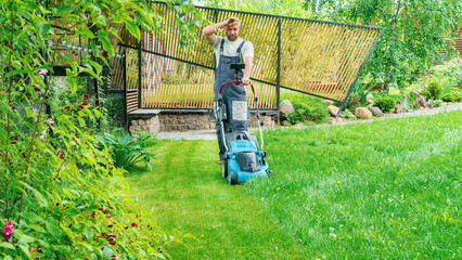 A man mows the lawn with an electric lawn mower in a landscaped garden. A man is tired from the tedious work of mowing the grass on the lawn on a hot summer day. Exhausting housework concept. - obrazy, fototapety, plakaty