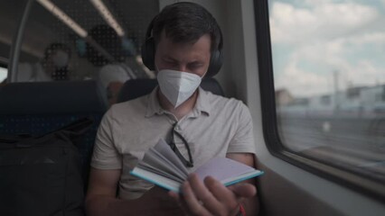 A male passenger rides a German suburban train in the summer wearing headphones and reading a book. Student reads textbook on wireless earphones during commute on train. Daily commute to education.  - Powered by Adobe