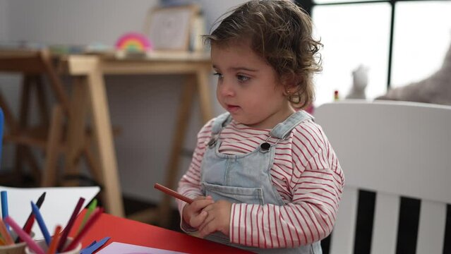 Adorable hispanic girl student sitting on table drawing on paper at kindergarten