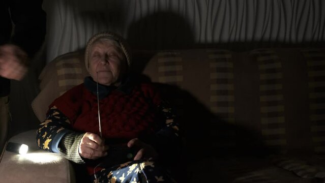 Grandmother holds radio in hands sitting in darkness blackout during russian terroristic attacks. Her family cover her legs by wrap and grandson hug. Family support concept. Together to the win.