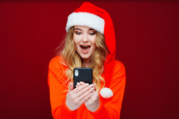 a surprised woman in a Santa Claus hat with smartphone on a red background. 