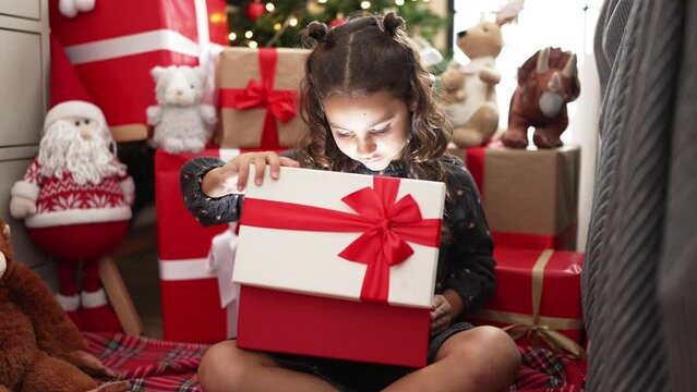 Adorable hispanic girl unpacking gift sitting on floor by christmas tree at home