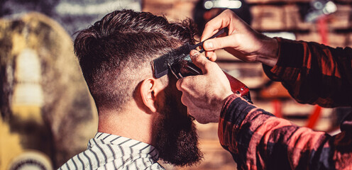 Hipster client getting haircut. Hands of barber with hair clipper, close up. Bearded man in...