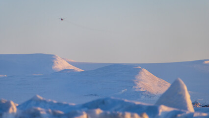 The helicopter is flying in the sky over the snow-capped mountains in the Far North in the Arctic. Passenger and cargo polar aviation. Northern landscape. Cold winter weather. Chukotka, Russia.